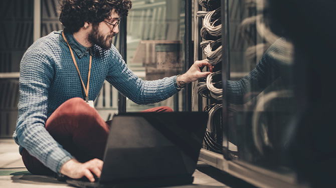 Ein IT-Fachmann sitzt mit seinem Notebook vor einem Serverschrank und überprüft Verbindungen.